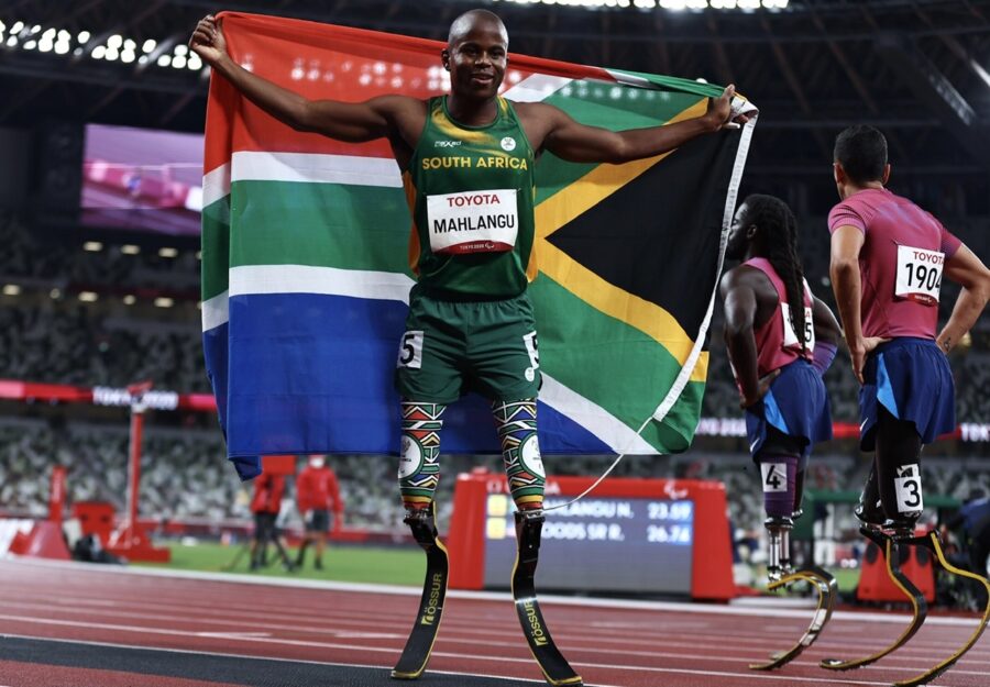 Tokyo 2020 Paralympic Games - Athletics - Men's 200m - T61 Final - Olympic Stadium, Tokyo, Japan - September 3, 2021. Ntando Mahlangu of South Africa celebrates after winning gold with the flag of South Africa whilst Regas Woods Sr of the United States and Luis Puertas of the United States look dejected REUTERS/Thomas Peter